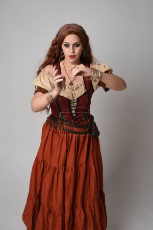 close up portrait of beautiful red haired woman wearing a medieval maiden, fortune teller costume. Posing with gestural hands reaching out, dancing, isolated on studio background.