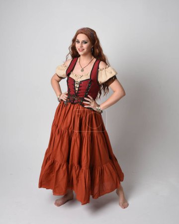 Photo for Full length portrait of beautiful red haired woman wearing a medieval maiden, fortune teller costume. Standing pose with dancing gestures, twirling skirt. isolated on studio background. - Royalty Free Image