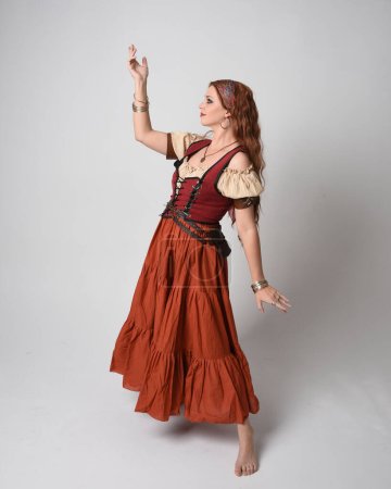 Full length portrait of beautiful red haired woman wearing a medieval maiden, fortune teller costume. Standing pose with dancing gestures, twirling skirt. isolated on studio background.