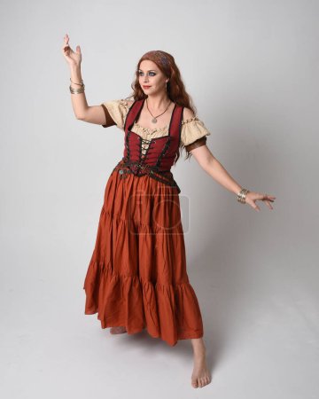 Photo for Full length portrait of beautiful red haired woman wearing a medieval maiden, fortune teller costume. Standing pose with dancing gestures, twirling skirt. isolated on studio background. - Royalty Free Image