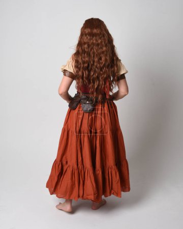 Portrait complet de belle femme aux cheveux roux portant une jeune fille médiévale, costume de diseuse de bonne aventure. Position debout vue de derrière, s'éloignant. isolé sur fond studio.