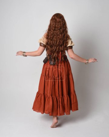 Téléchargez les photos : Portrait complet de belle femme aux cheveux roux portant une jeune fille médiévale, costume de diseuse de bonne aventure. Position debout vue de derrière, s'éloignant. isolé sur fond studio. - en image libre de droit