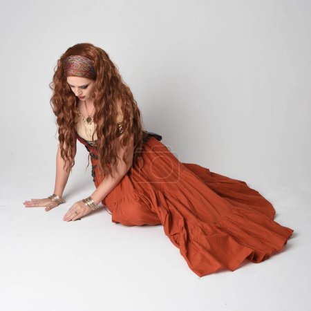 Portrait complet de belle femme aux cheveux roux portant une jeune fille médiévale, costume de diseuse de bonne aventure. Pose à genoux, assise par terre. isolé sur fond studio.