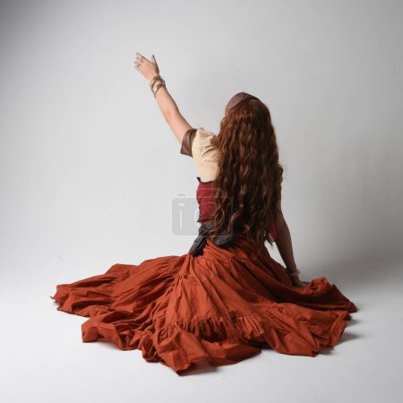 Téléchargez les photos : Portrait complet de belle femme aux cheveux roux portant une jeune fille médiévale, costume de diseuse de bonne aventure. Pose à genoux, assise par terre. isolé sur fond studio. - en image libre de droit