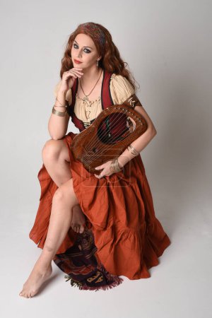 Portrait complet de belle femme aux cheveux roux portant une jeune fille médiévale, costume de diseuse de bonne aventure. Pose assise, instrument de lyre de musique. isolé sur fond studio.