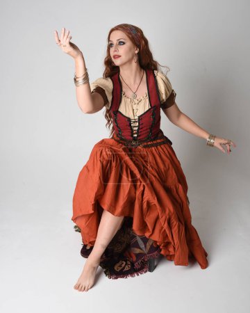 Ganztägiges Porträt einer schönen rothaarigen Frau, die ein mittelalterliches Mädchen- und Wahrsagerkostüm trägt. Sitzende Pose, gestikulierende Hände ausgestreckt. isoliert im Studio 