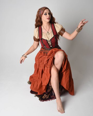 Ganztägiges Porträt einer schönen rothaarigen Frau, die ein mittelalterliches Mädchen- und Wahrsagerkostüm trägt. Sitzende Pose, gestikulierende Hände ausgestreckt. isoliert im Studio 