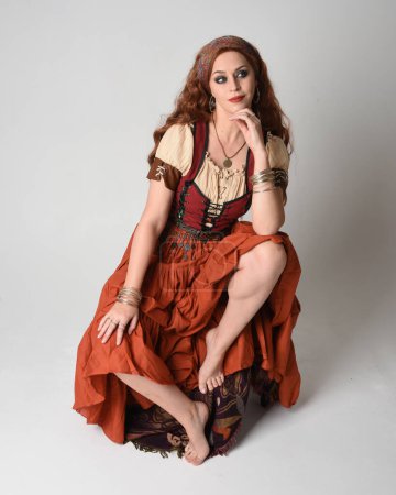 Photo for Full length portrait of beautiful red haired woman wearing a medieval maiden, fortune teller costume.  Sitting pose, with gestural hands reaching out. isolated on studio - Royalty Free Image