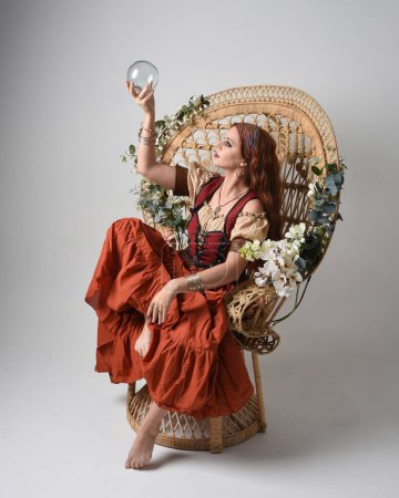 Foto de Retrato de cuerpo entero de una hermosa mujer pelirroja vestida con una doncella medieval, disfraz de adivina. Posar sentado, sosteniendo un orbe de cristal. aislado en el fondo del estudio. - Imagen libre de derechos
