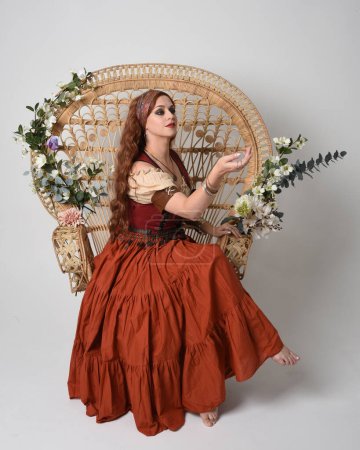 Retrato de cuerpo entero de una hermosa mujer pelirroja vestida con una doncella medieval, disfraz de adivina. Posar sentado, con las manos gestuales extendidas. aislado en el fondo del estudio.