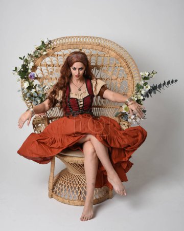 Portrait complet de belle femme aux cheveux roux portant une jeune fille médiévale, costume de diseuse de bonne aventure. Pose assise, les mains gestuelles tendues. isolé sur fond studio.
