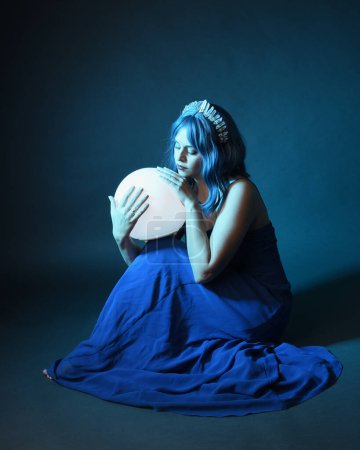 Photo for Full length portrait of beautiful female model wit blue hair wearing glamorous  fantasy ball gown with Crystal crown, holding a glowing orb lamp. Sitting pose, isolated on dark studio background - Royalty Free Image