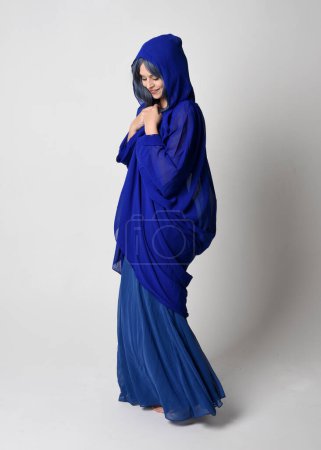 Photo for Full length portrait of beautiful female model wearing elegant fantasy blue ball gown, flowing cape with hood.Standing pose walking away, gestural arms reaching out. Isolated on  studio background - Royalty Free Image