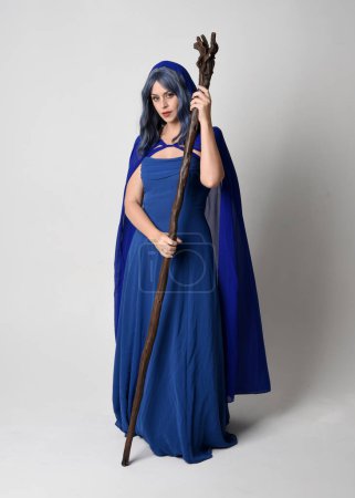 Full length portrait of beautiful female model wearing elegant fantasy blue ball gown, flowing cape with hood.Standing pose walking away, holding a wooden wizard staff. Isolated on white studio background.