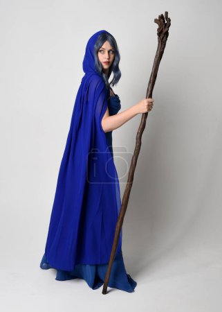 Full length portrait of beautiful female model wearing elegant fantasy blue ball gown, flowing cape with hood.Standing pose walking away, holding a wooden wizard staff. Isolated on white studio background.