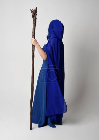 Photo for Full length portrait of beautiful female model wearing elegant fantasy blue ball gown, flowing cape with hood.Standing pose walking away, holding a wooden wizard staff. Isolated on white studio background. - Royalty Free Image
