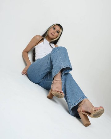 Photo for Full length portrait of brunette female asian model wearing casual clothes, white singlet shirt, denim jean pants. Sitting pose, high camera angle for perspective. Isolated on white studio background. - Royalty Free Image