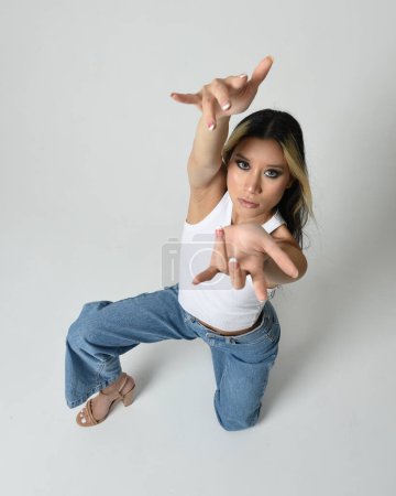 Photo for Full length portrait of brunette female asian model wearing casual clothes, white singlet shirt, denim jean pants. Sitting pose, high camera angle for perspective. Isolated on white studio background. - Royalty Free Image