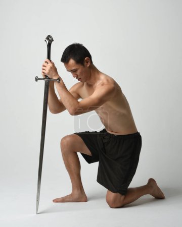 Photo for Full length portrait of fit handsome asian male model, wearing gym shorts and shirtless. Holding sword weapon,  kneeling in warrior training action pose, isolated on white studio background. - Royalty Free Image