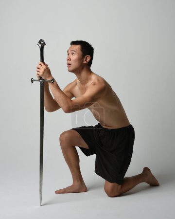 Photo for Full length portrait of fit handsome asian male model, wearing gym shorts and shirtless. Holding sword weapon,  kneeling in warrior training action pose, isolated on white studio background. - Royalty Free Image