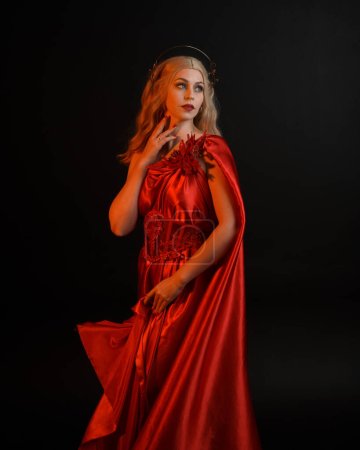Portrait en gros plan d'une belle mannequin blonde portant une toge de soie rouge fluide et une couronne, vêtue d'une ancienne déesse mythologique imaginaire. Gracieuse pose élégante isolée sur fond sombre studio.