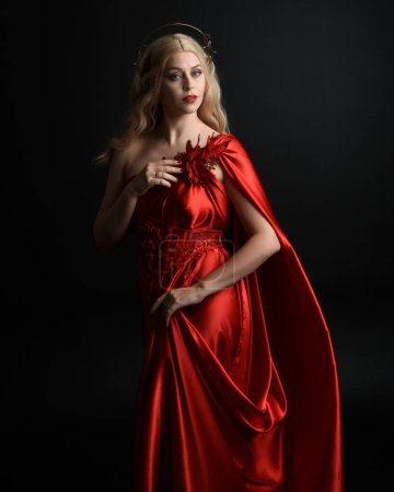 Portrait en gros plan d'une belle mannequin blonde portant une toge de soie rouge fluide et une couronne, vêtue d'une ancienne déesse mythologique imaginaire. Gracieuse pose élégante isolée sur fond sombre studio.