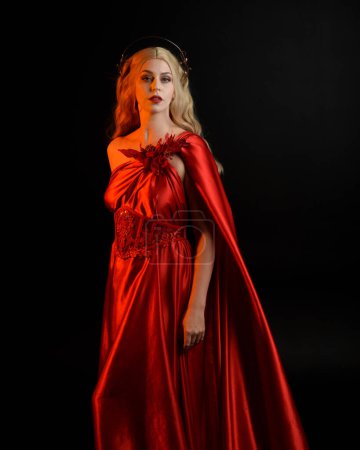 Nahaufnahme Porträt der schönen Blondine in fließendem roten Seiden-Toga-Kleid und Krone, gekleidet als antike mythologische Fantasie-Göttin. Anmutige elegante Pose isoliert auf dunklem Studiohintergrund.