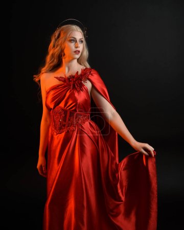 Nahaufnahme Porträt der schönen Blondine in fließendem roten Seiden-Toga-Kleid und Krone, gekleidet als antike mythologische Fantasie-Göttin. Anmutige elegante Pose isoliert auf dunklem Studiohintergrund.