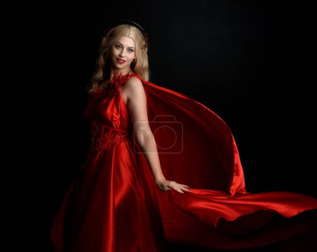 Primer plano retrato de la hermosa modelo rubia con vestido y corona de toga de seda roja que fluye, vestida como antigua diosa de la fantasía mitológica. Elegante pose elegante aislada sobre fondo oscuro del estudio.