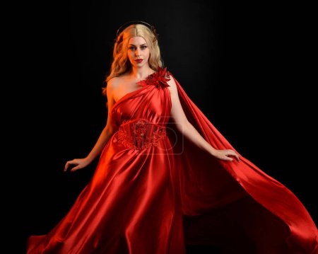 Primer plano retrato de la hermosa modelo rubia con vestido y corona de toga de seda roja que fluye, vestida como antigua diosa de la fantasía mitológica. Elegante pose elegante aislada sobre fondo oscuro del estudio.