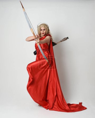 Full length portrait of beautiful blonde model dressed as ancient mythological fantasy goddess in flowing red silk toga gown, crown. Graceful elegant pose  holding  archery bow and arrow weapons, isolated on white studio background.