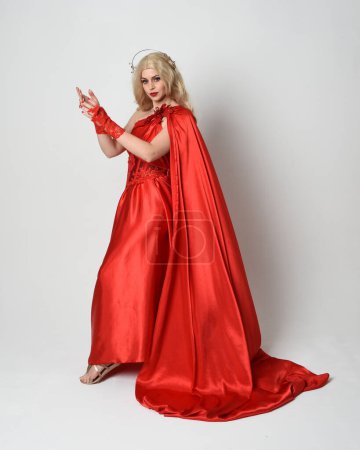Portrait de pleine longueur d'un beau modèle blond habillé en ancienne déesse mythologique fantaisie en robe de toge de soie rouge fluide, couronne. pose de danse élégante avec tissu fluide, isolé sur fond de studio blanc.