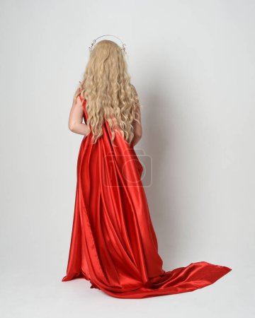 Photo for Full length portrait of beautiful blonde model dressed as ancient mythological fantasy goddess in flowing red silk toga gown, crown.   Walking away from camera,  isolated on white studio background. - Royalty Free Image