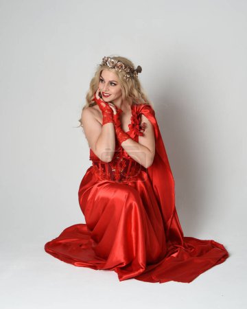Full length portrait of beautiful blonde model dressed as ancient mythological fantasy goddess in flowing red silk toga gown, crown. Kneeling pose sitting on floor. isolated on white studio background.