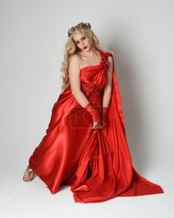 Photo for Full length portrait of beautiful blonde model dressed as ancient mythological fantasy goddess in flowing red silk toga gown, crown. Seated pose sitting on chair. isolated on white studio background. - Royalty Free Image