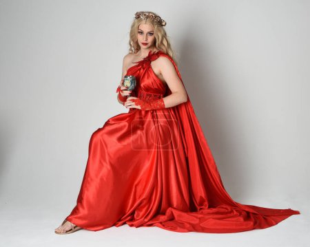 Full length portrait of beautiful blonde model dressed as ancient mythological fantasy goddess in flowing red silk toga gown, crown. Sitting pose holding wine goblet. isolated on white studio background.