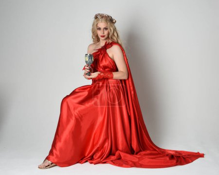 Photo for Full length portrait of beautiful blonde model dressed as ancient mythological fantasy goddess in flowing red silk toga gown, crown. Sitting pose holding wine goblet. isolated on white studio background. - Royalty Free Image