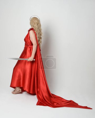 Full length portrait of beautiful blonde model dressed as ancient mythological fantasy goddess in flowing red silk toga gown, crown.  back view, walking away holding sword weapon,  isolated on white studio background.
