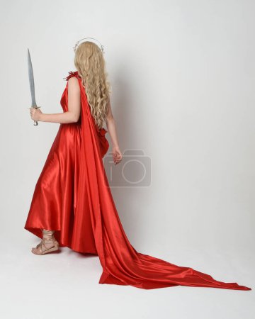 Photo for Full length portrait of beautiful blonde model dressed as ancient mythological fantasy goddess in flowing red silk toga gown, crown.  back view, walking away holding sword weapon,  isolated on white studio background. - Royalty Free Image
