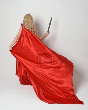 Portrait de pleine longueur d'un beau modèle blond habillé en ancienne déesse mythologique fantaisie en robe de toge de soie rouge fluide, couronne. vue de dos, marchant loin tenant arme épée, isolé sur fond de studio blanc.