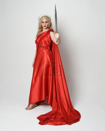 Ganzkörperporträt der schönen blonden Modell als antike mythologische Fantasie Göttin in fließenden roten Seide Toga Kleid, Krone gekleidet. Gehen in Pose, eine Schwertwaffe in der Hand, isolierter Studiohintergrund