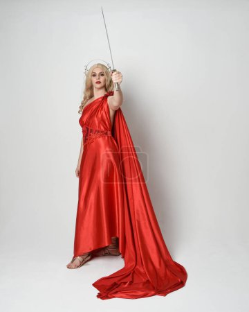 Full length portrait of beautiful blonde model dressed as ancient mythological fantasy goddess in flowing red silk toga gown, crown.   walking pose,  holding a sword weapon, isolated studio background