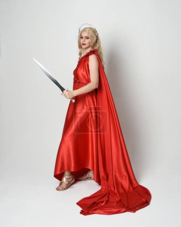 Photo for Full length portrait of beautiful blonde model dressed as ancient mythological fantasy goddess in flowing red silk toga gown, crown.   walking pose,  holding a sword weapon, isolated studio background - Royalty Free Image