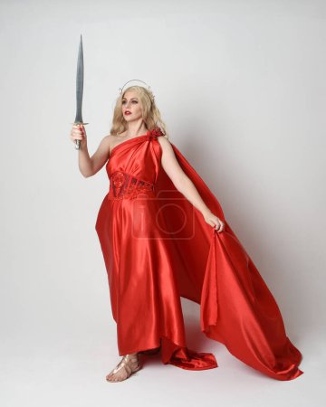 Photo for Full length portrait of beautiful blonde model dressed as ancient mythological fantasy goddess in flowing red silk toga gown, crown.   walking pose,  holding a sword weapon, isolated studio background - Royalty Free Image