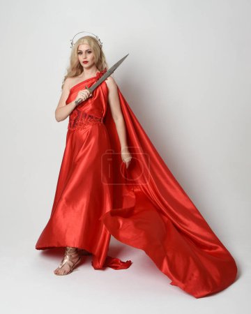 Ganzkörperporträt der schönen blonden Modell als antike mythologische Fantasie Göttin in fließenden roten Seide Toga Kleid, Krone gekleidet. Gehen in Pose, eine Schwertwaffe in der Hand, isolierter Studiohintergrund