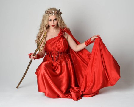 Full length portrait of beautiful blonde model dressed as ancient mythological fantasy goddess in flowing red silk toga gown, crown. kneeling pose,  golden trident weapon, isolated studio background