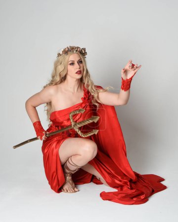 Ganzkörperporträt der schönen blonden Modell als antike mythologische Fantasie Göttin in fließenden roten Seide Toga Kleid, Krone gekleidet. kniende Pose, goldene Dreizack-Waffe, isolierter Studiohintergrund