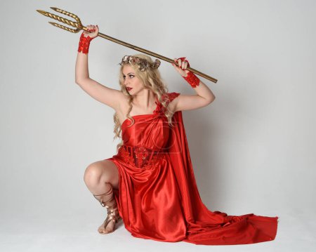 Portrait de pleine longueur d'un beau modèle blond habillé en ancienne déesse mythologique fantaisie en robe de toge de soie rouge fluide, couronne. pose à genoux, arme trident dorée, fond studio isolé