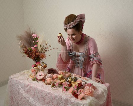Primer plano retrato de modelo femenina linda con un vestido rosa opulento, el traje de una nobleza barroca francesa histórica. Comer alimentos y dulces en una fiesta indulgente en la mesa cargada de pasteles, flores y joyas ricas.