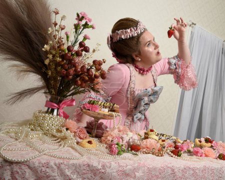 Gros plan portrait de mignonne mannequin féminine portant une robe rose opulente, le costume d'une noblesse baroque française historique. Manger des repas gourmands avec des pâtisseries, des fleurs et de riches bijoux